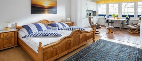 Quarto com cama de madeira, tapete azul estampado, pintura de um pôr do sol e vista para uma cozinha e área de jantar.