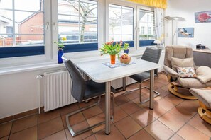 A room with a dining table, chairs, a radiator, a window with curtains, and a chair with a cushion. - Holiday apartment in Norderney with Wi-Fi (Norderney)