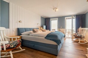 Un dormitorio con una cama, una mecedora, una mesita de noche y una ventana con cortinas.
