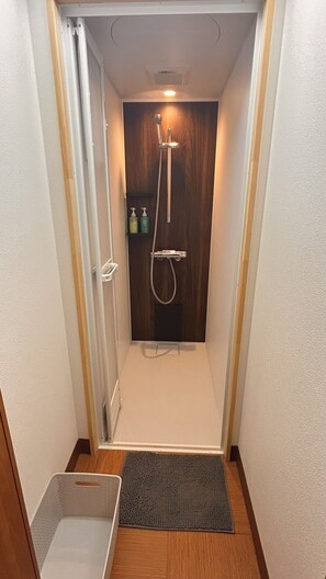 Ein schmales Duschbad mit Holztür, Duschbrause und einem kleinen Ablagefach für Toilettenartikel.