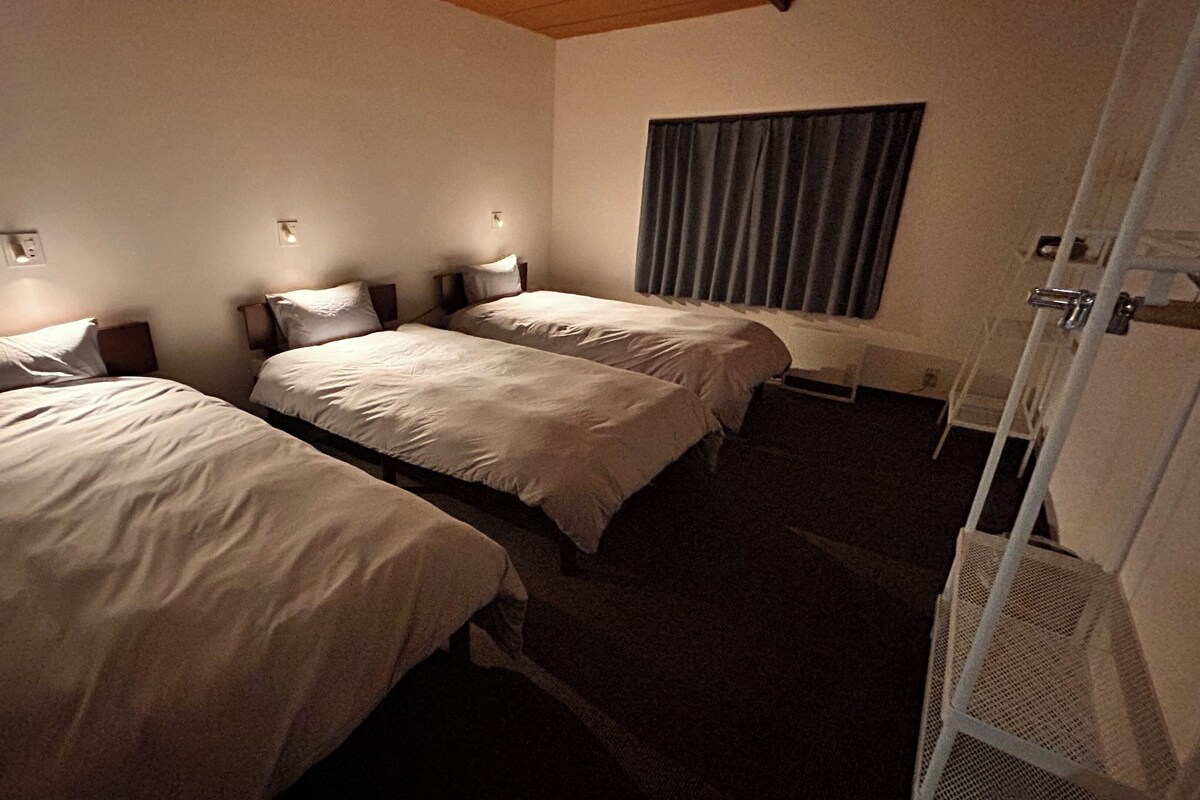 Ein Hotelzimmer mit zwei Einzelbetten, einem Fenster mit Vorhängen, einem Heizkörper und einer Tür mit Metallgriff.