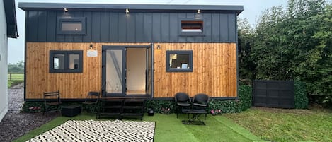 A modern house with a wooden exterior, a sliding door entrance, and a small patio with two chairs and a patterned rug.