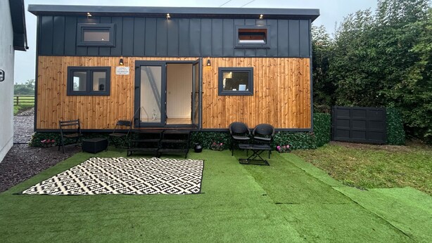 A modern, compact building with a wooden exterior, a glass door entrance, and a small outdoor seating area with chairs and a table. - Cabin - Sleeps 6 - Parking (Meath)