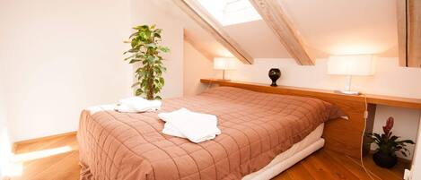 A bedroom with a sloped ceiling, a bed with a brown quilted bedspread, two bedside lamps, a potted plant, and a wooden nightstand.