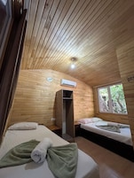 Una habitación con paneles de madera, una cama con toalla verde, una mesita de noche, un ventanal con vistas a árboles y un equipo de aire acondicionado.