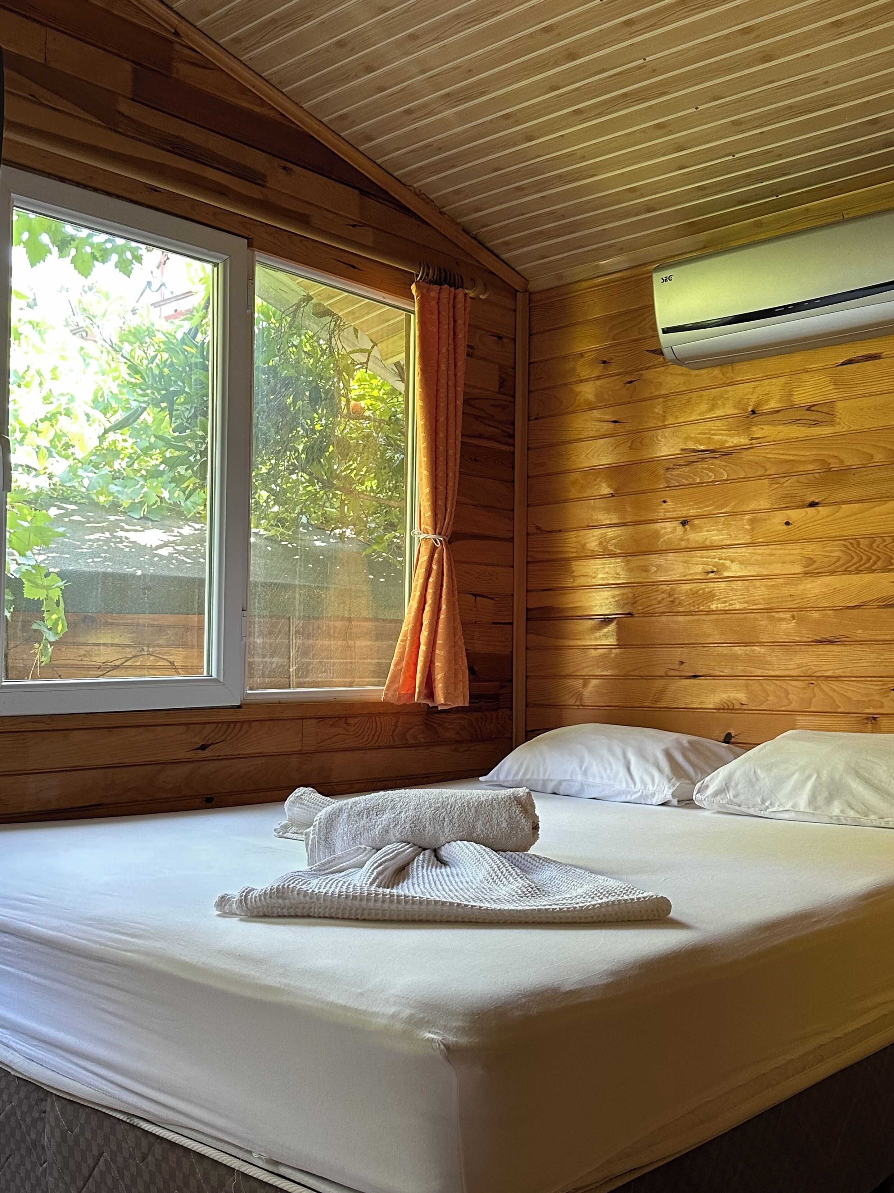 A wooden cabin room with a bed, a window with curtains, and an air conditioning unit.