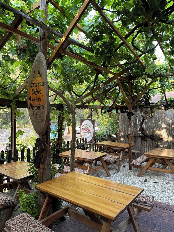 An outdoor seating area with wooden tables and benches, surrounded by greenery and a wooden pergola.