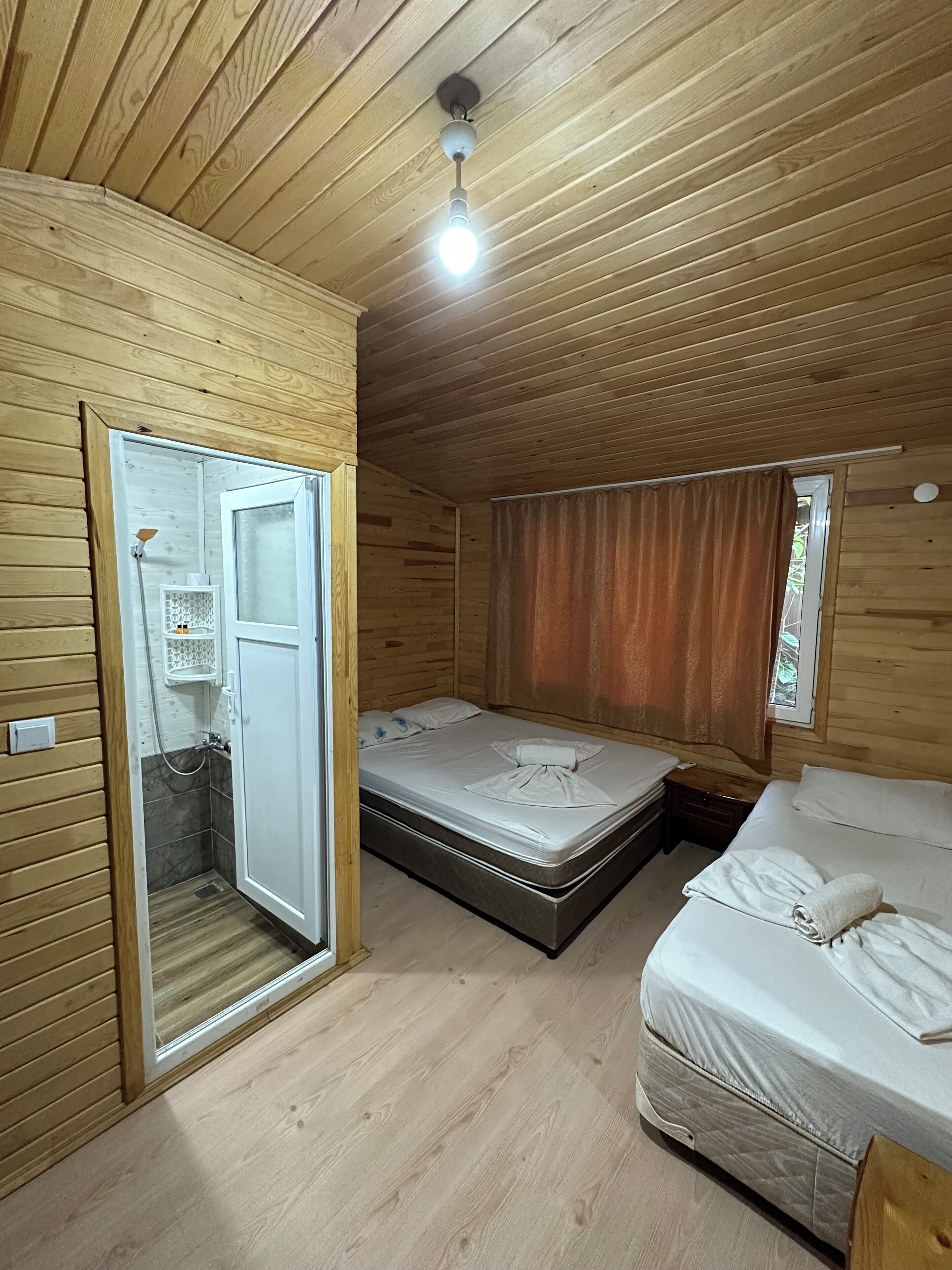 A room with a wooden ceiling, a bed with white bedding, a nightstand, and a bathroom with a shower.