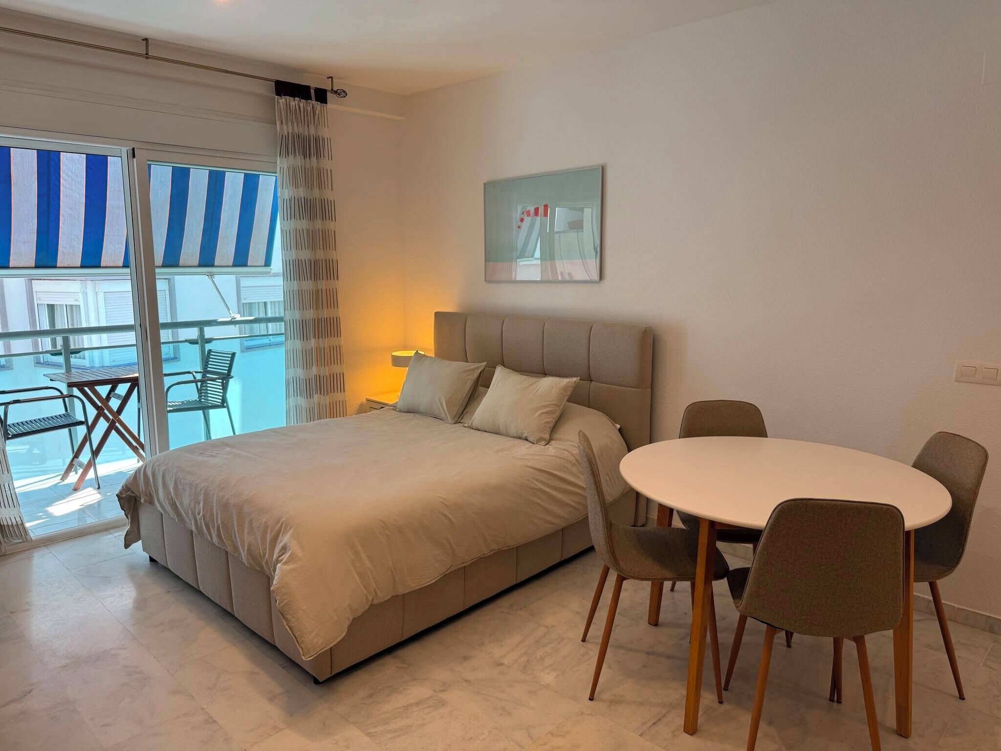 A hotel room with a bed, a dining table with chairs, a balcony with a striped awning, and a painting on the wall.