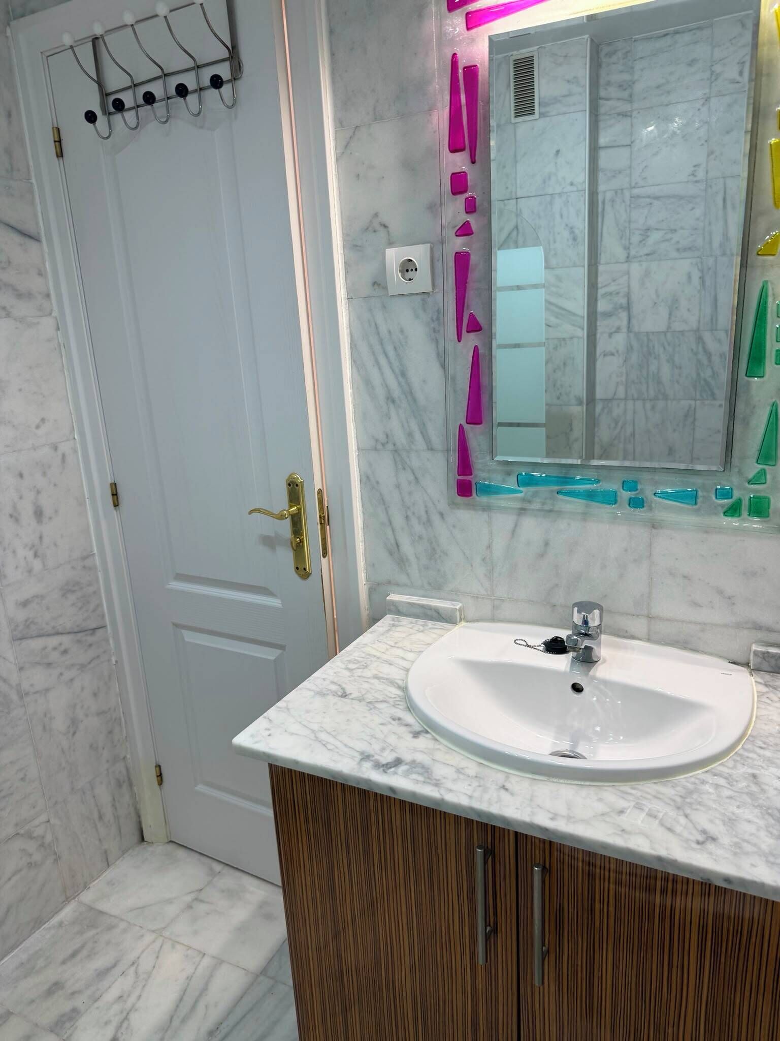 A bathroom with a white marble sink, wooden vanity, and a mirror with colorful glass decorations.