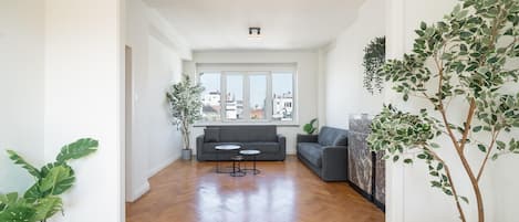 A spacious living area with a wooden floor, a grey sofa, and two potted plants.