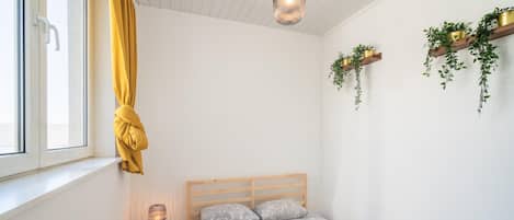 A bedroom with a bed, a bedside table with a lamp, a window with curtains, and wall-mounted shelves with plants.