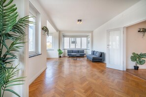 Une pièce spacieuse avec un plancher en bois, un canapé gris et une petite table. De grandes fenêtres laissent entrer la lumière naturelle et offrent une vue sur l’extérieur.