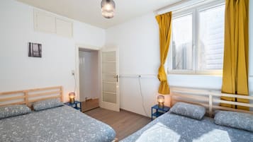 Une chambre avec deux lits, une fenêtre avec des rideaux jaunes, un luminaire suspendu et une porte donnant sur une autre pièce.