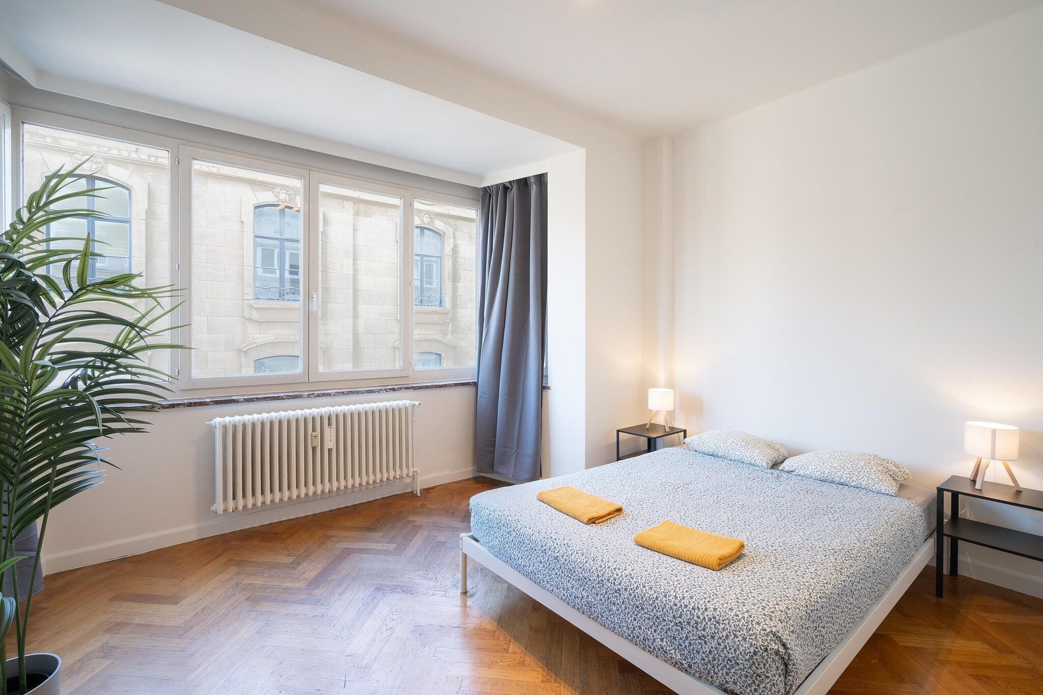A bedroom with a bed, two bedside lamps, a radiator, and a window with curtains.