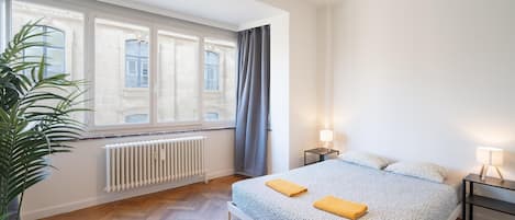 A bedroom with a bed, two bedside lamps, a radiator, and a window with curtains.
