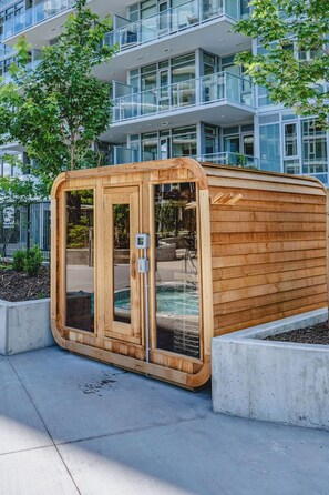 Estructura de sauna de madera con puertas de vidrio, ubicada sobre una superficie de concreto y con una piscina visible a través de las puertas.