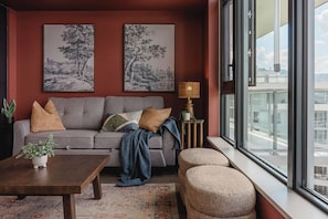 A modern living room with a sofa, coffee table, and ottomans, featuring artwork on the walls and a view through large windows. - The Southwest Pottery Suite | DT Kelowna 1BR | Pool&Sauna (Kelowna)