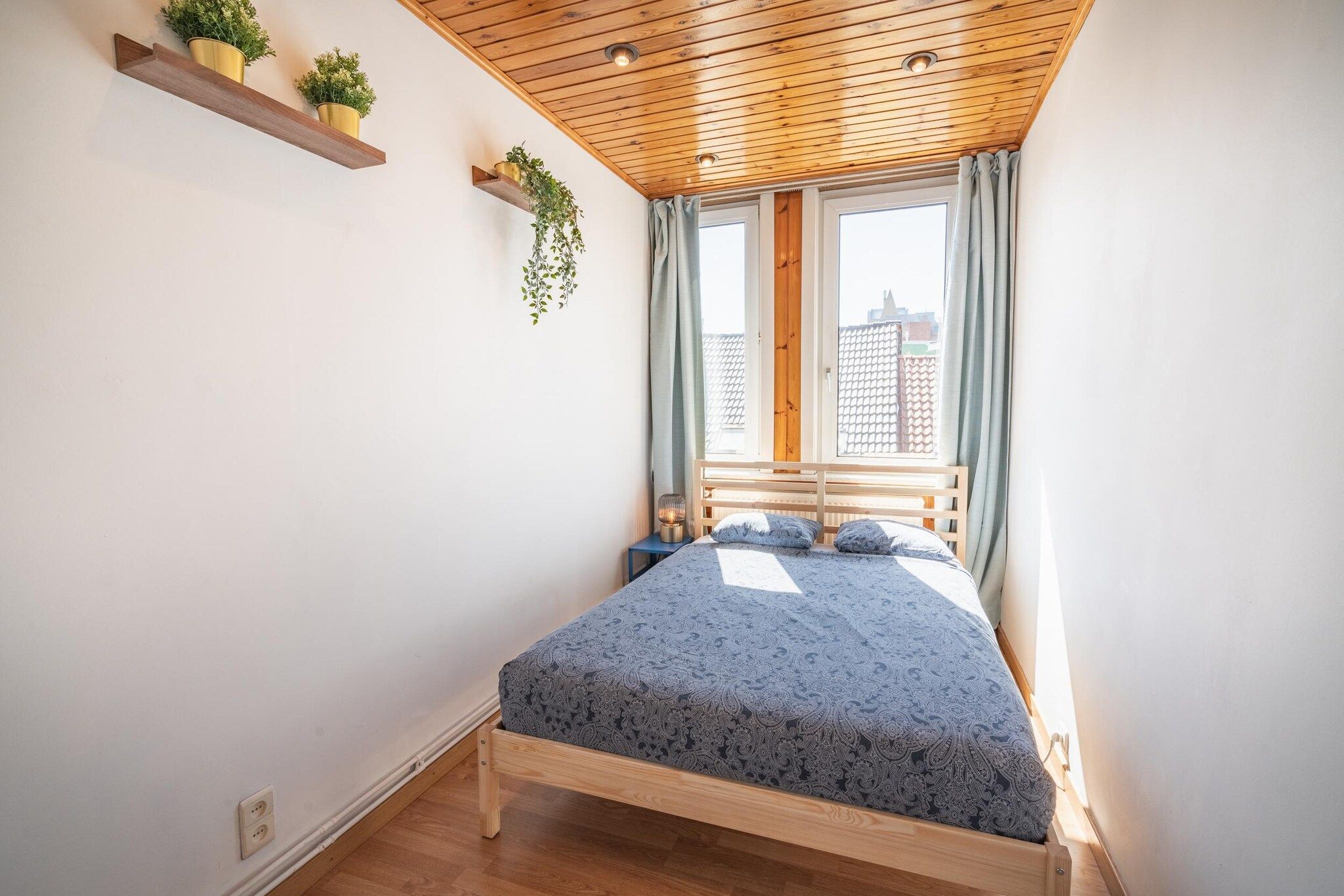 Une chambre avec un plafond en bois, un lit recouvert d’une couvre-lit à motifs, une fenêtre avec des rideaux et deux étagères murales ornées de plantes.