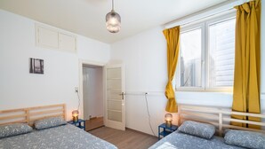 Un dormitorio con dos camas, una ventana con cortinas amarillas, una lámpara colgante y una puerta que da a otra habitación.