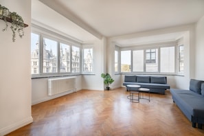 A spacious living room with large windows, a radiator, a grey sofa, a small round table, and a potted plant. - Family-Friendly 3-Bedroom Home (Antwerp)