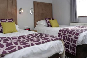 Ein Hotelzimmer mit zwei Betten, jeweils mit einem gemusterten Überwurf, einem Nachttischchen mit einer Uhr und einer Wandleuchte.