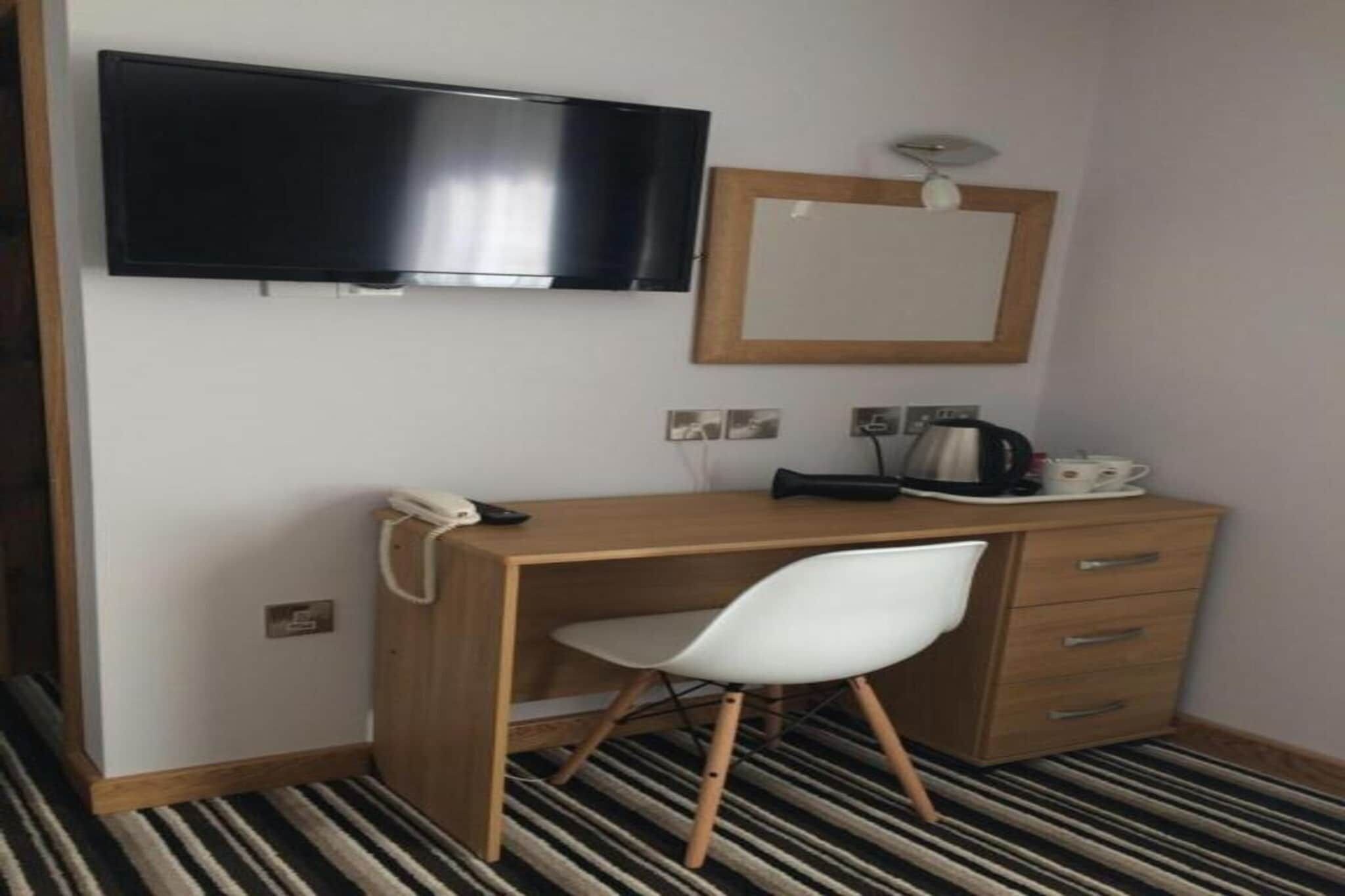 A hotel room with a wooden desk, a white chair, a flat-screen TV mounted on the wall, a small mirror, a telephone, and a kettle on the counter.