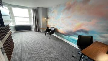A hotel room with a large window, a carpeted floor, a desk, a chair, and a wall with a painted sky mural.