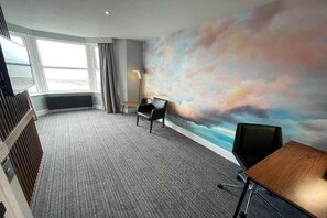 Une chambre d’hôtel dotée d’une grande fenêtre, d’un sol recouvert de moquette, d’un bureau, d’une chaise et d’un mur orné d’une fresque représentant un ciel peint.