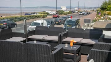 Outdoor wicker furniture with cushions, a table with a drink, and a view of a busy road and water body.