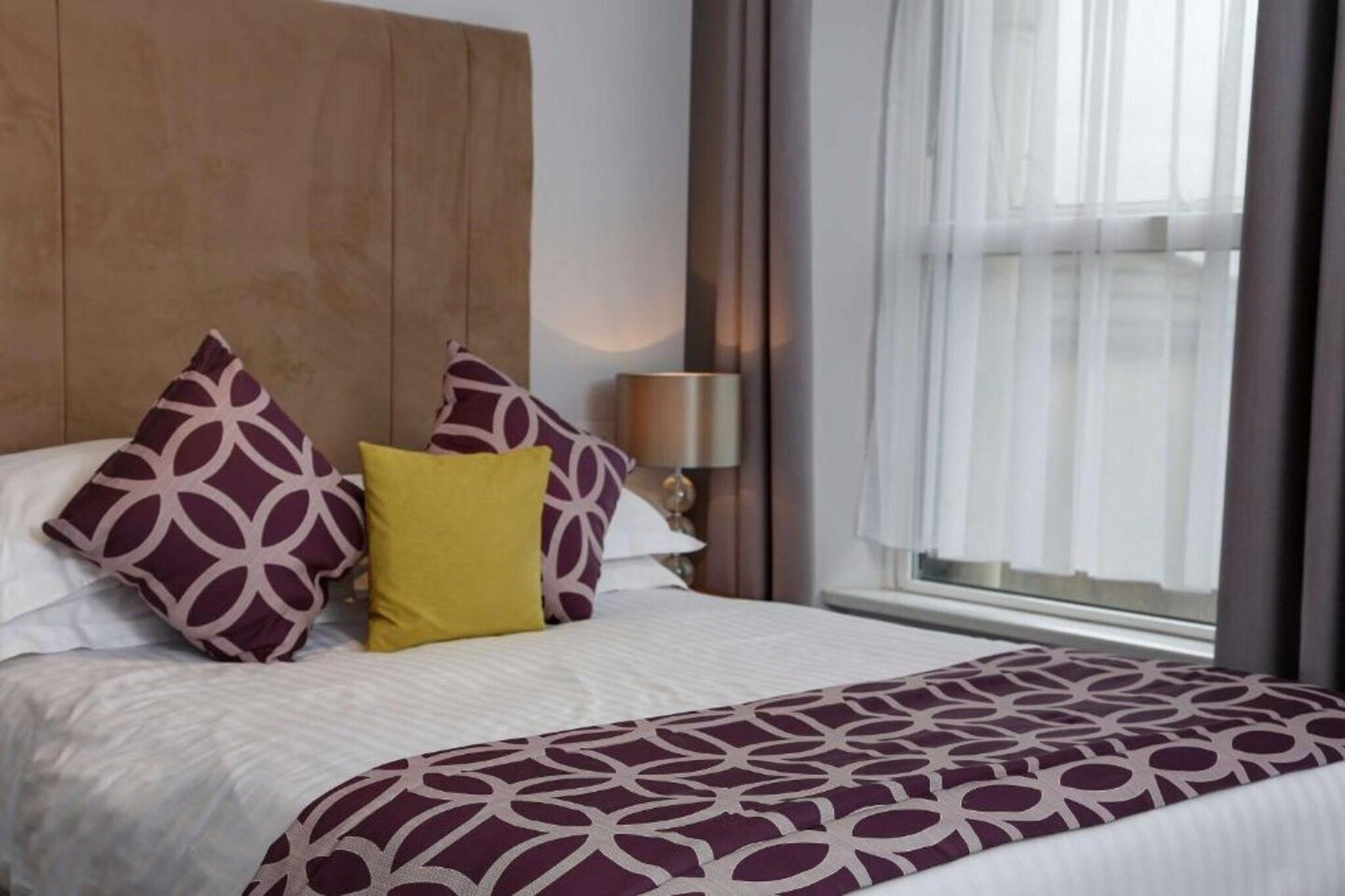 A hotel room with a large bed, a wooden headboard, a window with curtains, and a bedside lamp.