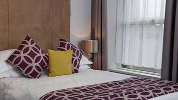 A hotel room with a large bed, a wooden headboard, a window with curtains, and a bedside lamp.