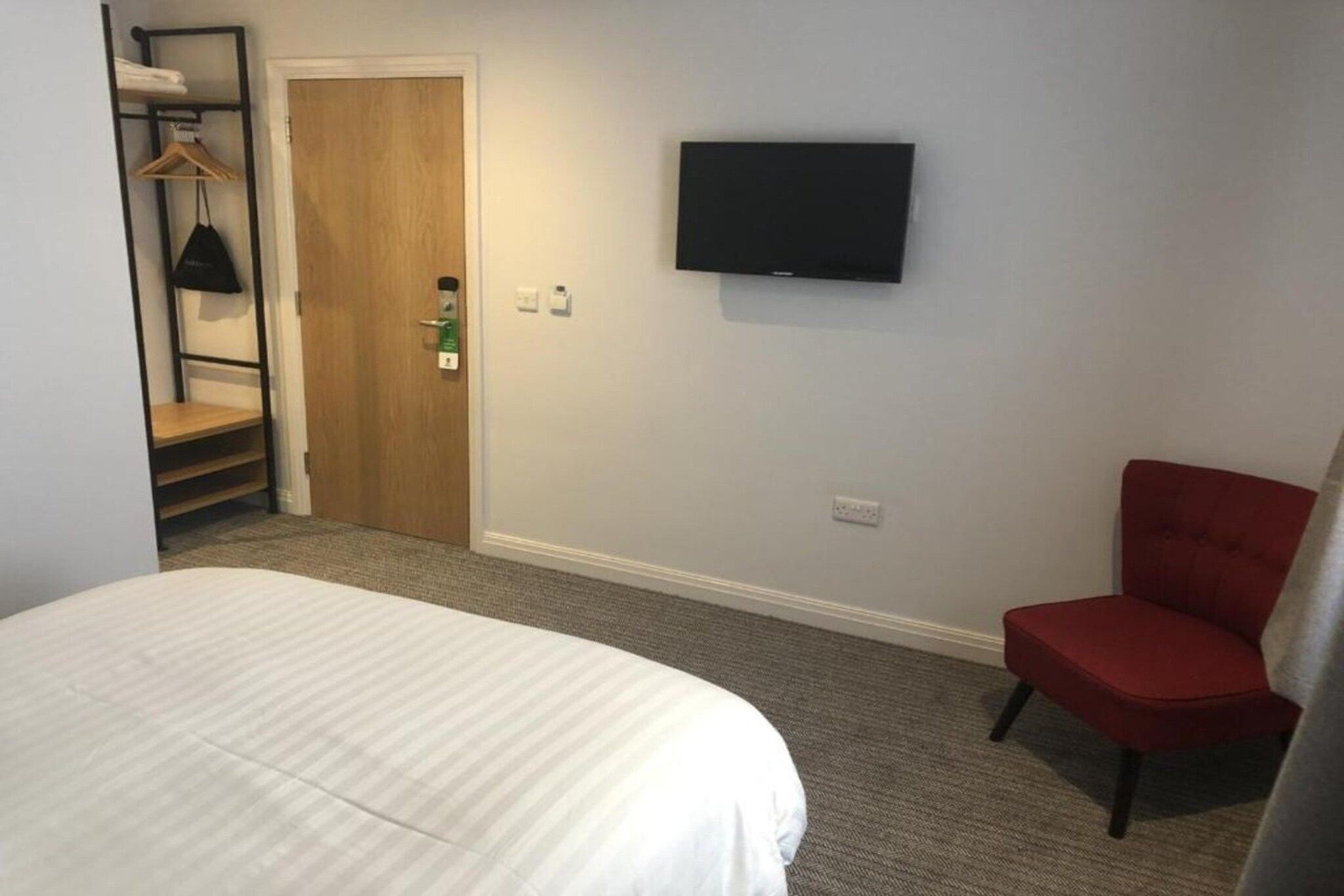 A hotel room with a bed, a red chair, a television mounted on the wall, and a wooden door.
