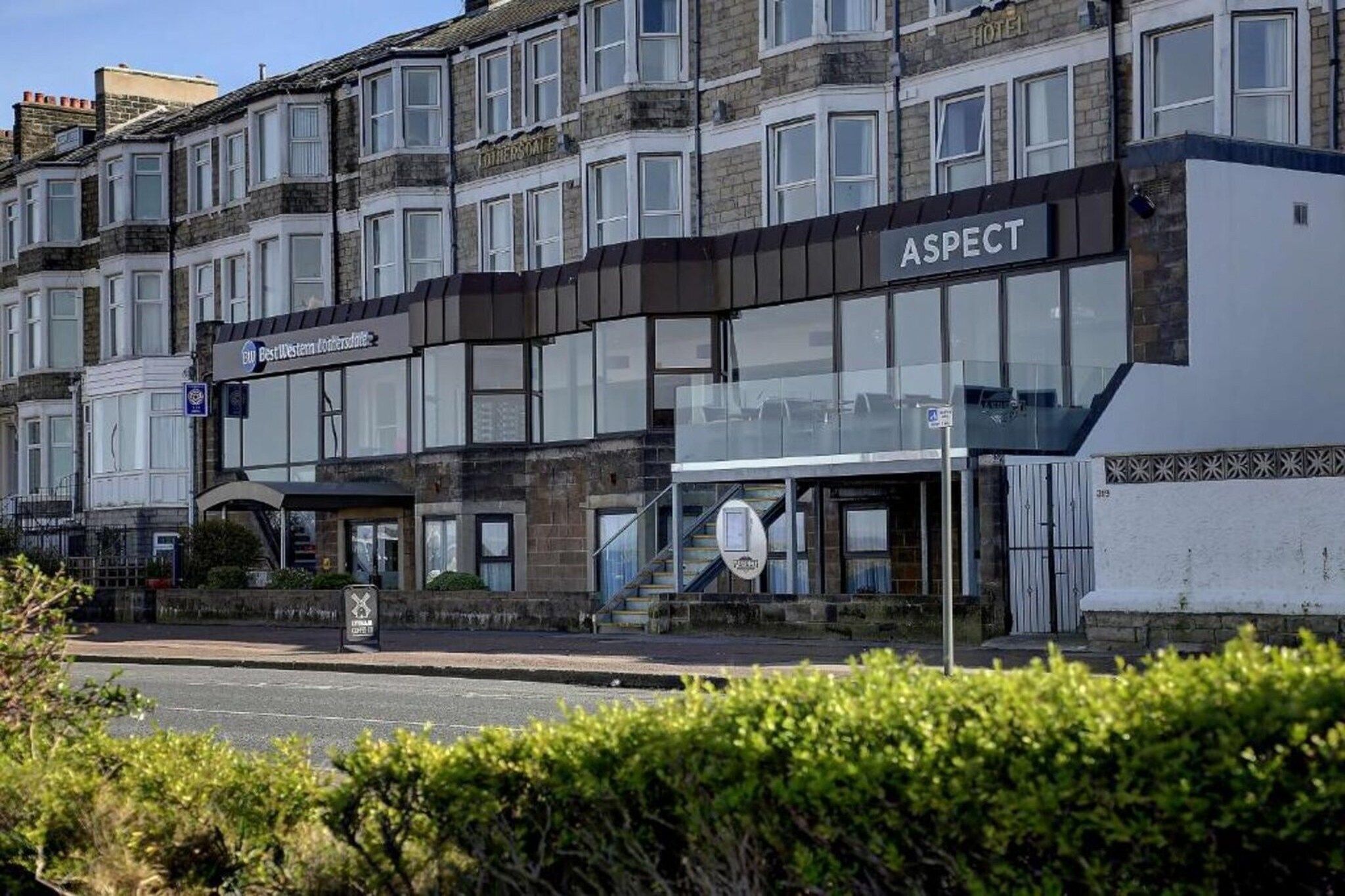 A hotel named ASPECT with a modern facade, a visible entrance, and a sign indicating a nearby parking area.