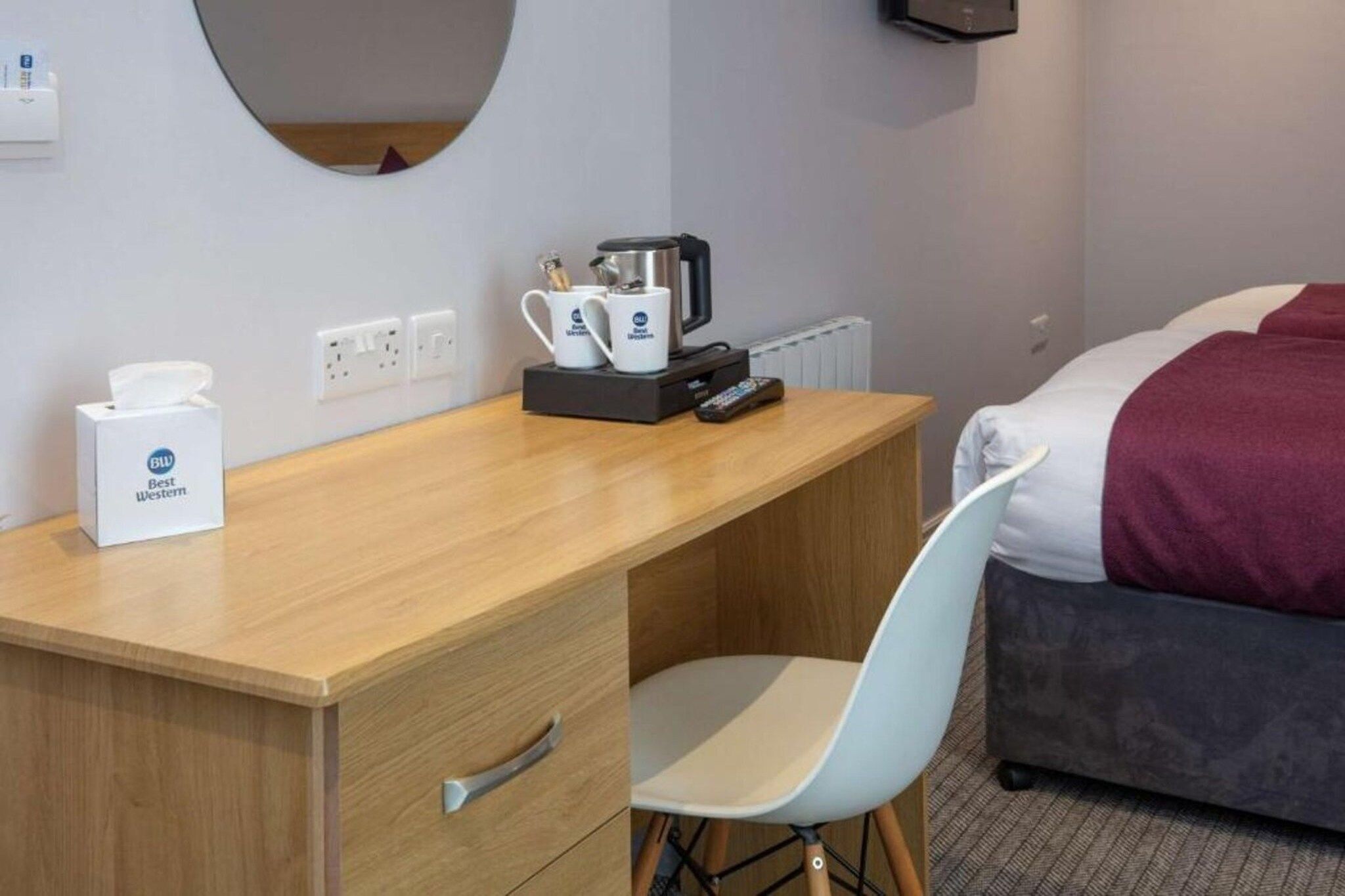 A hotel room with a wooden desk, a chair, a bed with a red blanket, a coffee maker, and a tissue box.