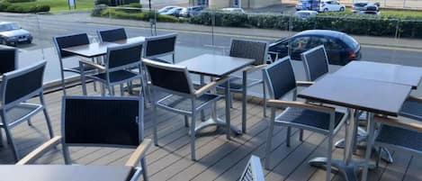 An outdoor seating area with tables and chairs, a view of the sea, and a building in the background.