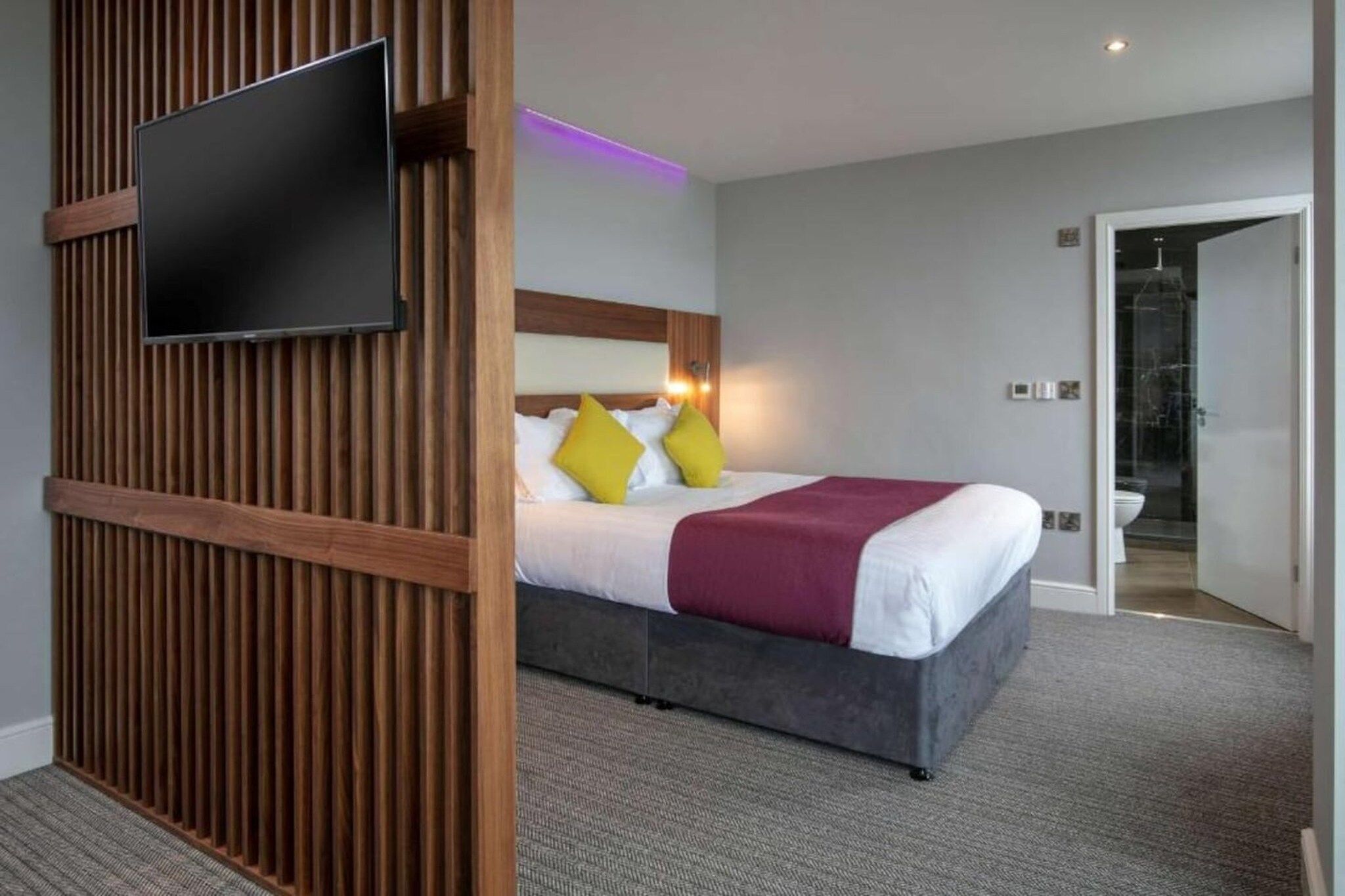 A hotel room with a bed, a television mounted on a wooden panel, and a bathroom visible through an open door.
