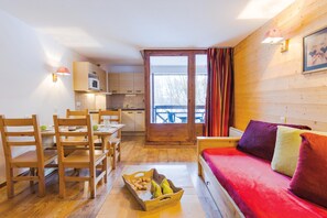 Ein gemütliches Zimmer mit Holzboden, einem roten Sofa, einem Esstisch mit Stühlen, einer Küchenzeile und einem großen Fenster mit Vorhängen.