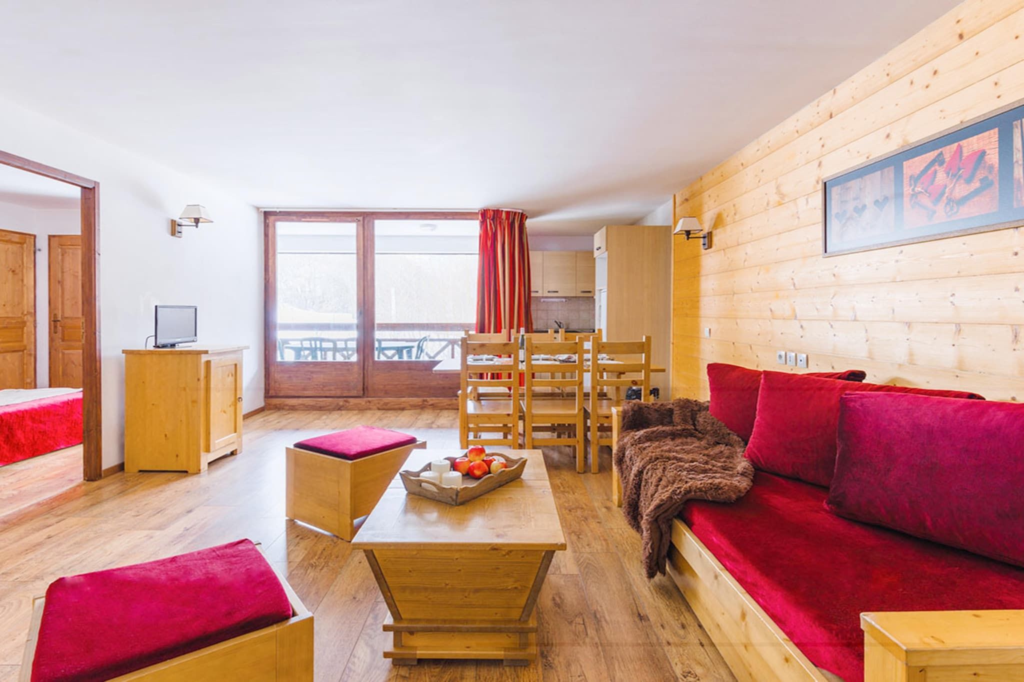 A cozy room with a wooden floor, a red sofa, a wooden table with a fruit bowl, and a wooden chair.