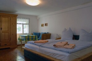 Un dormitorio con una cama doble, un armario de madera, una mesita con mantel a cuadros y una ventana con cortina blanca.
