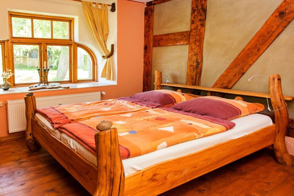 A wooden bed in a room with a large window, a radiator, and a wooden ceiling beam.