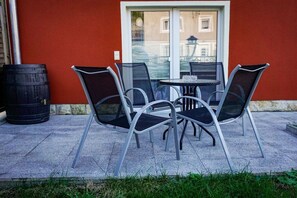 Eine Terrasse mit Tisch und vier Stühlen, einem schwarzen Fass und einer roten Wand mit einem Fenster.