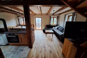 Une cuisine avec des meubles en bois et un fourneau, un salon avec un canapé et une télévision, et une grande fenêtre qui laisse entrer la lumière naturelle.