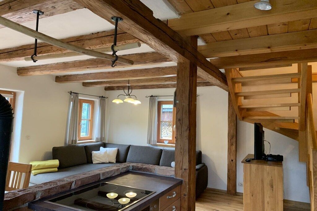 A rustic interior with exposed wooden beams, a stone fireplace, a seating area with cushions, and a kitchen with a built-in stove.