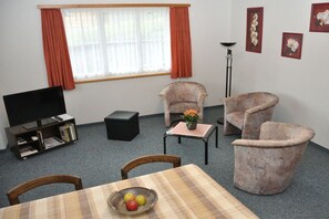 Una sala de estar con televisor, sofá, sillón, mesa con fruta y ventana con cortinas.