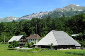 Ländliche Szenerie mit zwei traditionellen Holzhäusern, eines davon mit Balkon, umgeben von üppiger Vegetation und Bergen im Hintergrund.
