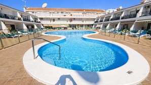 Piscina de borda curva, rodeada por espreguiçadeiras e um edifício com varandas.