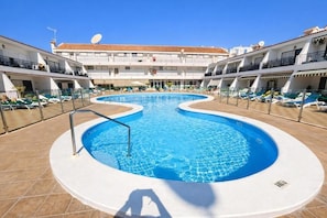 A swimming pool with a curved edge, surrounded by lounge chairs and a building with balconies. - Apartment Chipeque (Arona)