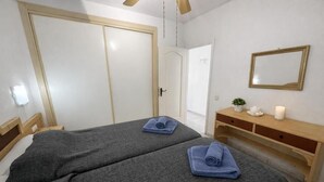 Quarto de hotel com duas camas, uma cômoda de madeira, um espelho e uma porta que dá para outro espaço.