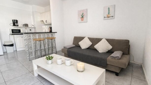 Uma sala de estar moderna com um sofá, uma mesa de centro e uma cozinha ao fundo.
